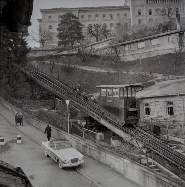 Ein historisches Foto zeigt das Bundeshaus in Bern, ein grosses mehrstöckiges Gebäude mit Bogenfenstern. Es steht auf einer Anhöhe, umgeben von Bäumen und Wegen. Die Drahtseilbahn führt vom angelegten Garten nach oben.Ein Fall für Humoristen
